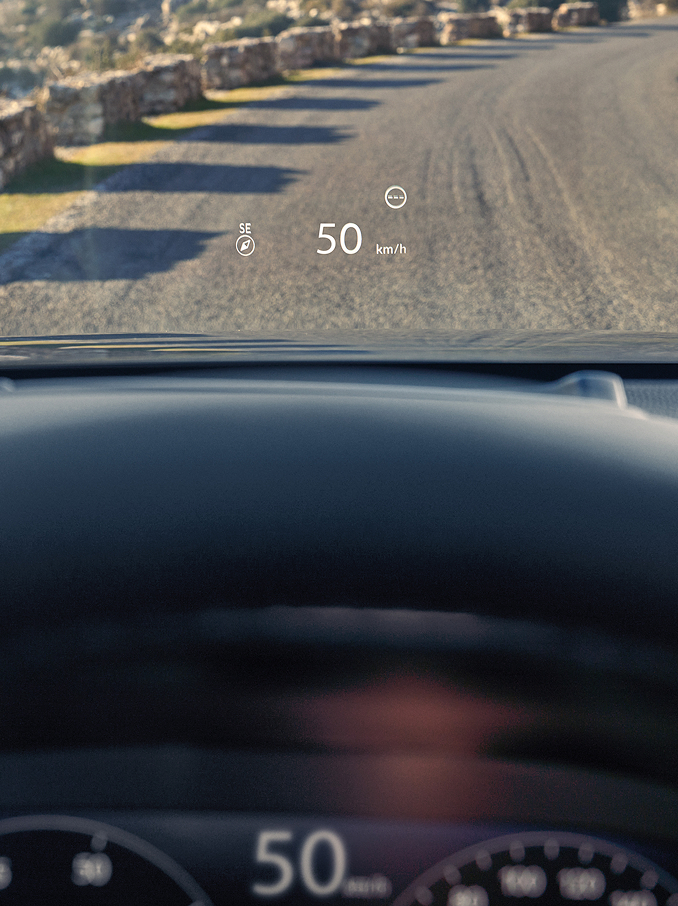 Una visuale in prima persona dal sedile del conducente mostra un Head-Up Display (HUD) che proietta "50 km/h" e una bussola sul parabrezza sopra una strada sterrata.