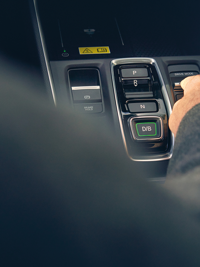 La mano di una persona si allunga per regolare un interruttore a levetta sulla console centrale di un'auto moderna, accanto ai pulsanti del selettore del cambio.