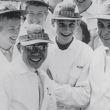 Soichiro Honda e un gruppo di operai che indossano una tuta da lavoro bianca.
