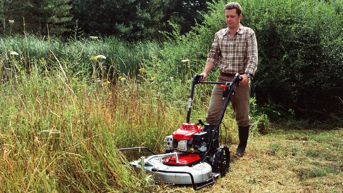Inquadratura frontale di tre quarti di un rasaerba professionale rivolto verso sinistra utilizzato da un operatore. Luogo di utilizzo: giardino.