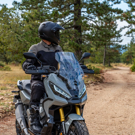 Honda X-ADV, vista anteriore di tre quarti destra, guidatore in piedi su moto grigia, sentiero nella foresta