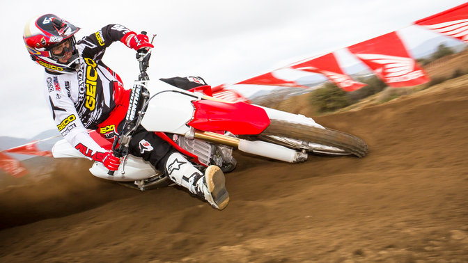 Inquadratura frontale di tre quarti della CRF250R con il pilota piegato in curva. Strada sterrata.