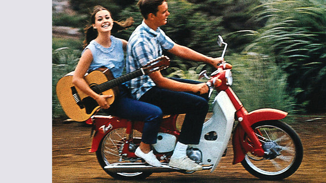 Motorino Honda con pilota e passeggera con chitarra. Strada polverosa. Lato destro.