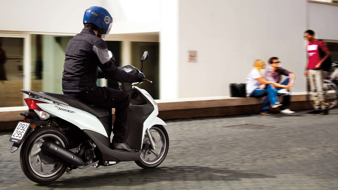 Inquadratura posteriore di tre quarti dello scooter Honda Vision 50 bianco con pilota. Lato destro (su strada).