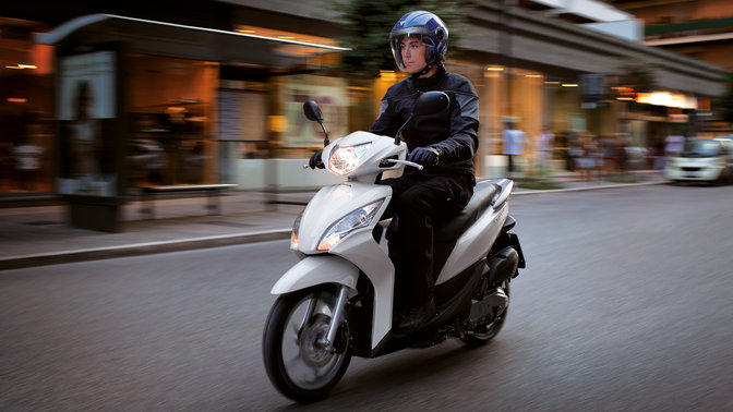 Inquadratura anteriore di tre quarti dello scooter Honda Vision bianco con pilota. Lato sinistro (su strada).