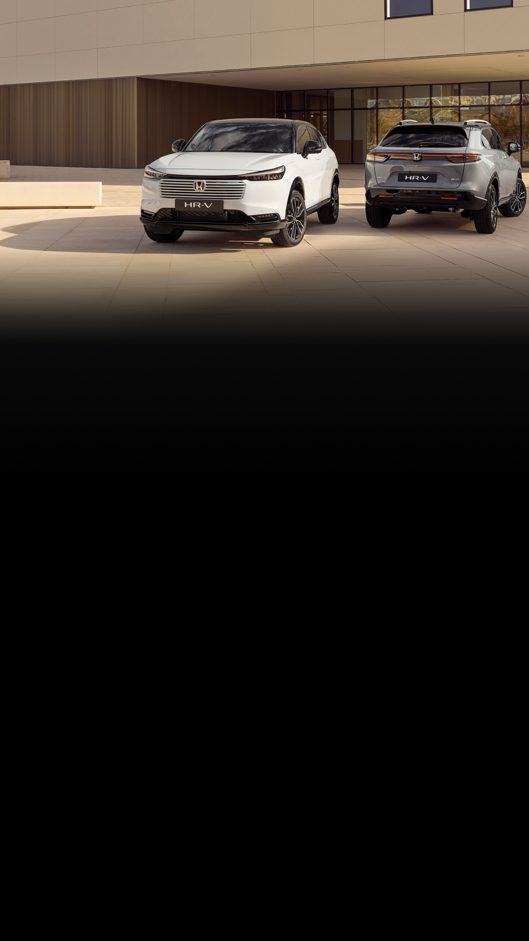 Two Honda HR-V SUVs parked in front of a modern building. The white HR-V is in the foreground facing forward, while the silver HR-V is in the background facing away, showcasing the front and rear designs of the vehicle.