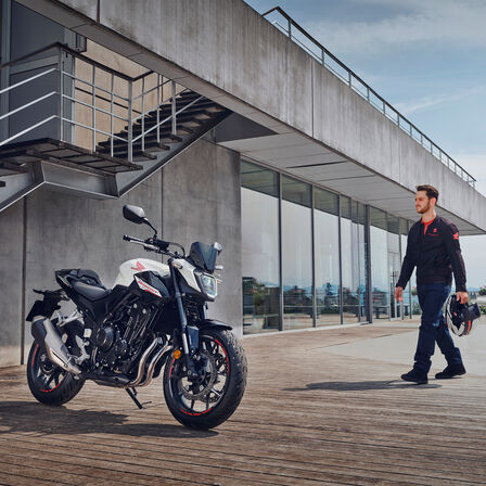 L'immagine mostra un uomo che cammina verso una Honda CB500 Hornet parcheggiata in un ambiente urbano moderno. La moto è visibile con un design aggressivo e sportivo, mentre l'uomo si prepara a indossare il casco, suggerendo l'inizio di un'avventura su strada.