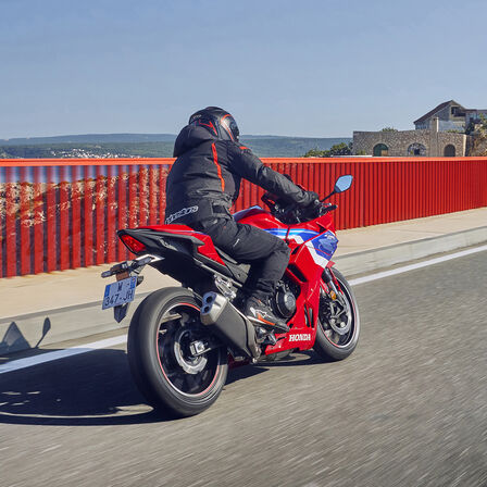 L'immagine mostra un motociclista in sella a una Honda CBR500R, mentre percorre una strada in un paesaggio estivo. Il design sportivo della moto, abbinato all'abbigliamento da corsa del pilota, evoca una sensazione di velocità e performance. La scena si svolge su una strada panoramica, suggerendo un'esperienza di guida entusiastica e dinamica.